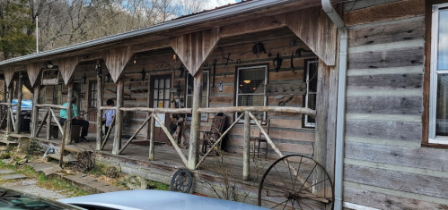A rustic wooden cabin with a porch, featuring old tools and decorations, surrounded by trees and a gravel path.