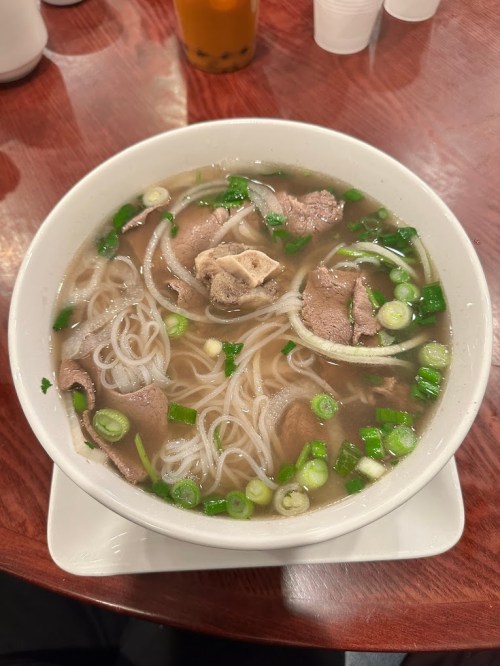 A bowl of pho with broth, rice noodles, sliced meat, and green onions, served on a wooden table.