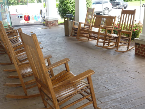 A row of wooden rocking chairs on a porch, with a view of a parking area and colorful decorations in the background.