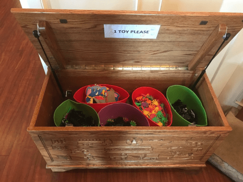 A wooden toy chest with an open lid, containing colorful bins filled with various toys. A sign reads "1 Toy Please."