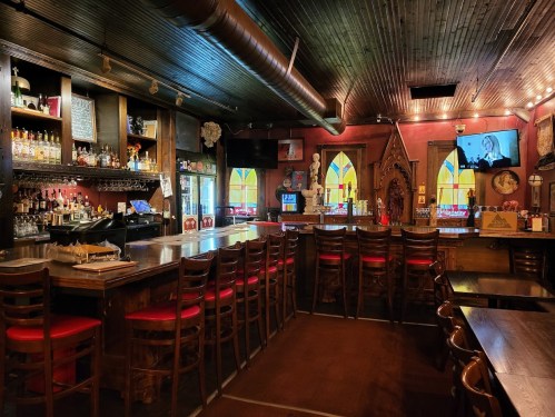 A cozy bar interior with wooden accents, a long counter, red chairs, and colorful stained glass windows.