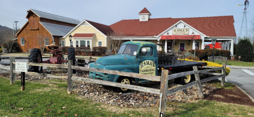 A vintage blue truck parked in front of a farmhouse-style restaurant with a red roof and a tractor nearby.