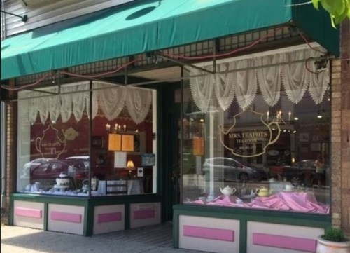 A charming tea shop with a green awning, lace curtains, and pink accents, showcasing teapots and tea sets in the window.