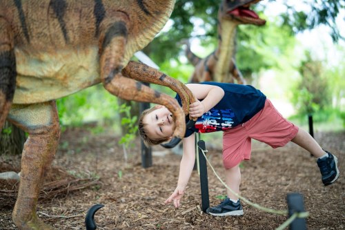 A child playfully interacts with a dinosaur statue in a lush outdoor setting.