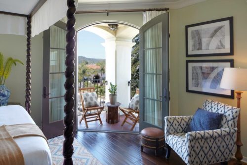 A cozy hotel room with open doors leading to a balcony, featuring two chairs and a view of greenery and mountains.