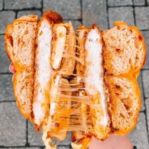 A close-up of a gooey, cheesy sandwich with layers of bread, melted cheese, and crispy filling, held in a hand.