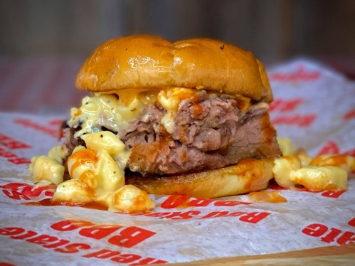 A close-up of a juicy burger with pulled pork, melted cheese, and macaroni on a toasted bun, served on paper.