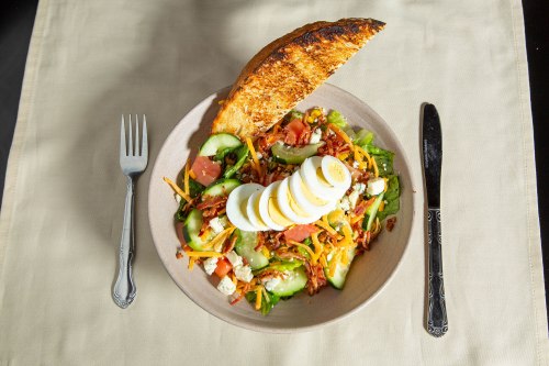 A colorful salad with sliced eggs, tomatoes, cucumbers, and cheese, served with a toasted bread slice on a beige tablecloth.