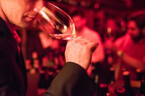 A person in a suit holds a wine glass, examining it in a dimly lit room filled with bottles.