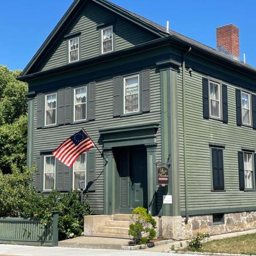 A green historic house with black shutters and an American flag displayed in front, under a clear blue sky.