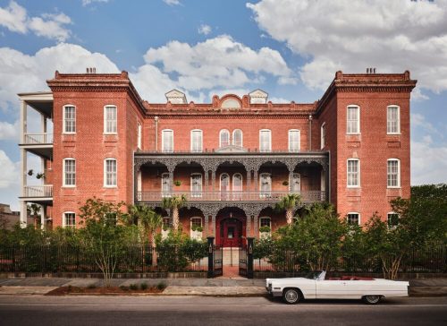 Historic red brick building with ornate balconies, surrounded by greenery, and a classic white convertible parked in front.
