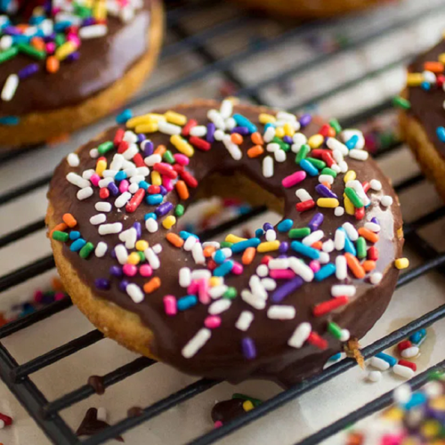 A chocolate frosted donut topped with colorful sprinkles on a cooling rack, surrounded by scattered sprinkles.