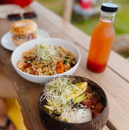 A wooden table with a bowl of rice topped with avocado and sprouts, a plate of fried rice, and a bottle of drink.