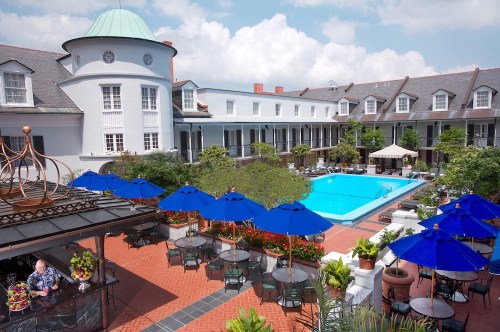 A sunny courtyard with a pool, surrounded by greenery and blue umbrellas, next to a charming hotel building.