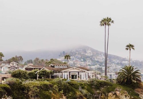 Coastal view featuring palm trees, houses on a hillside, and foggy mountains in the background.