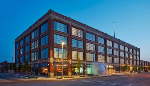 A large brick building with many windows, illuminated at dusk, located on a city street.