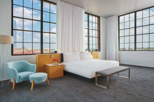 Bright, modern hotel room with large windows, a white bed, a blue chair, and minimalist decor.