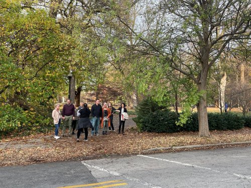 A group of people gathered in a park with trees and fallen leaves, chatting near a lamp post.