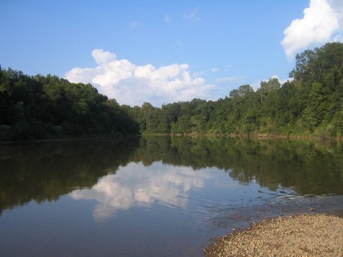 A calm river reflects blue skies and fluffy clouds, surrounded by lush green trees and a pebbly shore.
