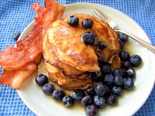 A plate of fluffy pancakes topped with blueberries, served with crispy bacon on a blue textured background.