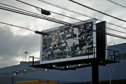 A weathered billboard with peeling paint and faded text, set against a cloudy sky and power lines.