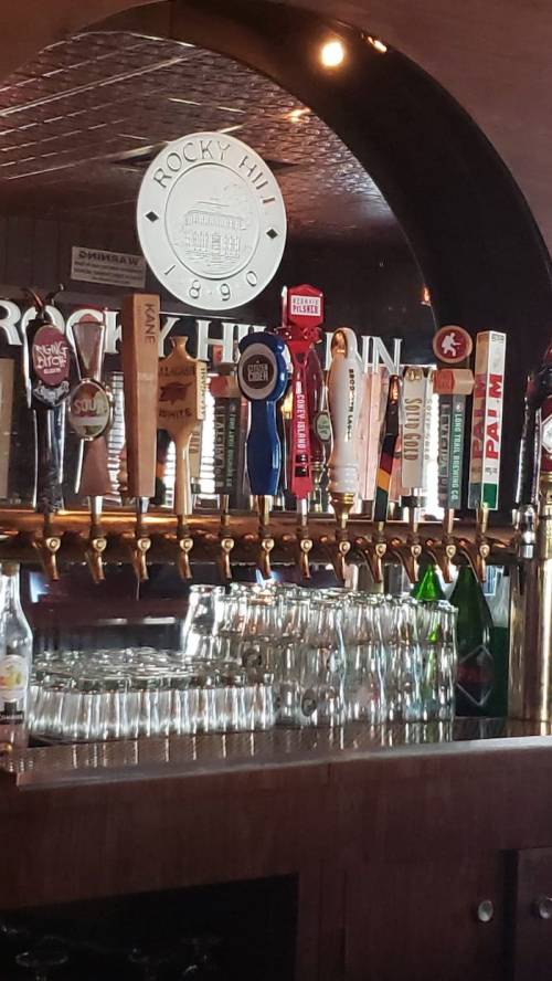 A bar with various beer taps and a mirror displaying "Rocky Inn 1890" in the background. Glasses are stacked below.
