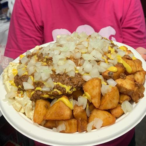 A large plate of loaded fries topped with chili, cheese, and diced onions, held by a person in a pink shirt.
