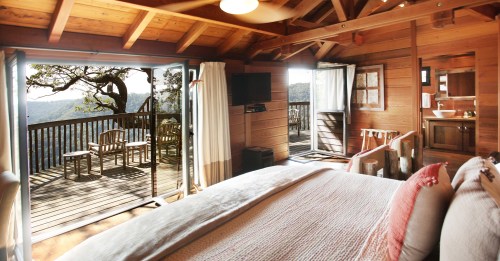 Cozy wooden cabin interior with a bed, balcony view, and natural light streaming in from large windows.