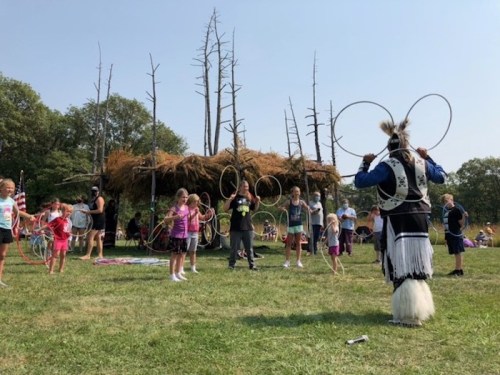 A group of children and adults participate in a hoop dance workshop outdoors, with a traditional structure in the background.