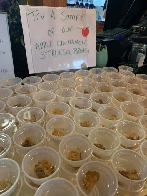 A table with numerous small cups containing samples of apple cinnamon streusel bread, with a sign inviting people to try.