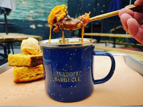 A hand holds a chopstick with cheesy barbecue meat above a blue mug, accompanied by cornbread on a tray.