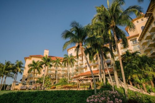 A tropical resort with palm trees, lush greenery, and a sunny sky, featuring a multi-story building.