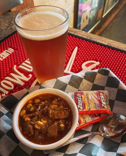 A bowl of hearty chili with corn, a spoon, and two seasoning packets, alongside a glass of amber beer.