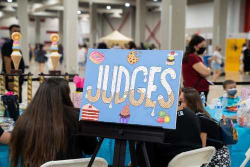 A colorful sign reading "JUDGES" on an easel, with people seated at a table in a busy event space.