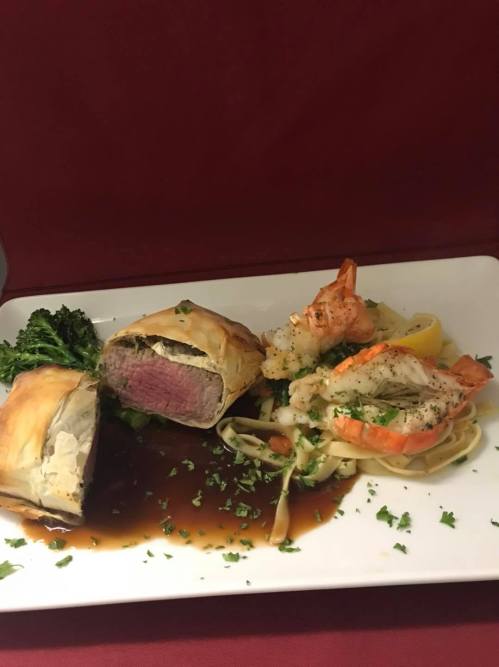 A gourmet plate featuring beef wellington, shrimp, and fettuccine, garnished with herbs on a white dish.