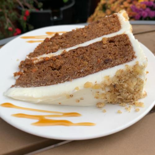 A slice of carrot cake with cream cheese frosting, topped with chopped nuts and drizzled with caramel sauce.
