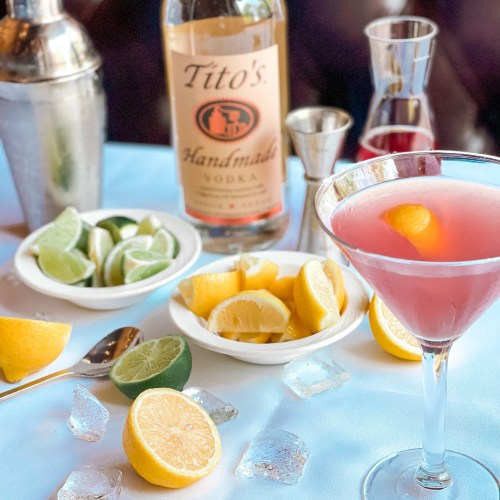 A pink cocktail garnished with an orange slice, surrounded by lemons, limes, ice, and a bottle of Tito's vodka.