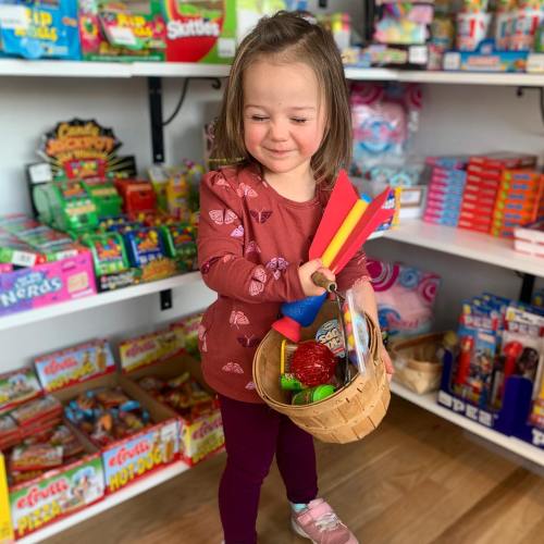 A young girl smiles while holding a basket of colorful toys in a bright, cheerful store filled with various products.