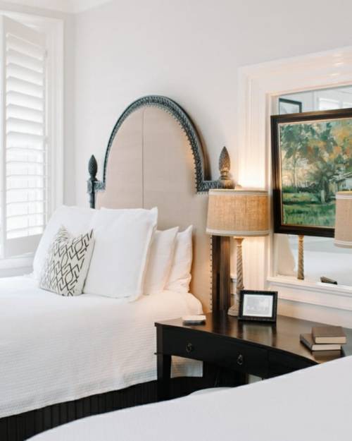 A bright bedroom featuring two beds, a decorative headboard, lamps, and a mirror reflecting a painting.
