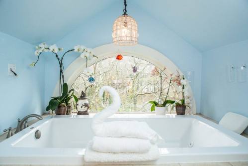 A serene bathroom with a blue ceiling, a swan-shaped towel, and plants by a large arched window.