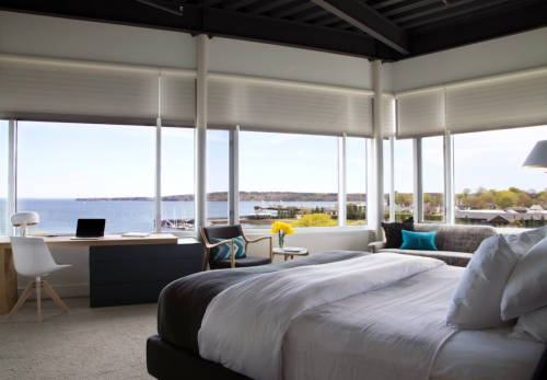 Bright, modern bedroom with large windows overlooking a scenic waterfront view and cozy furnishings.