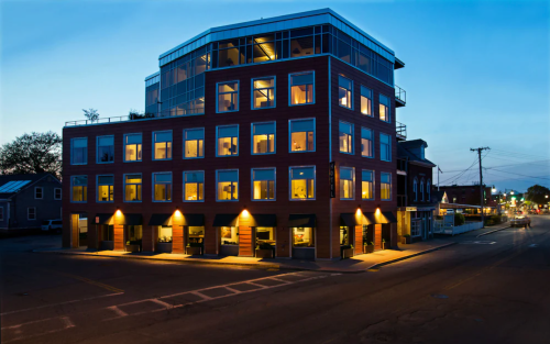 Modern building with large windows, illuminated at dusk, located at a street corner.