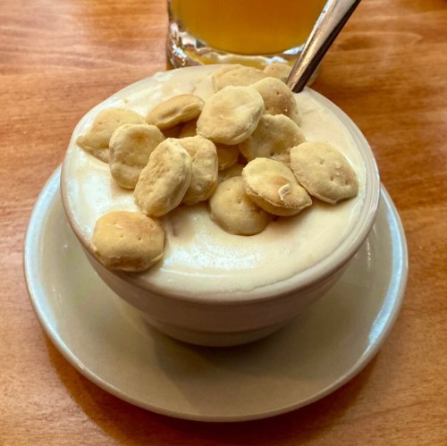 A small bowl of creamy dessert topped with crunchy snacks, placed on a wooden table next to a glass of beverage.