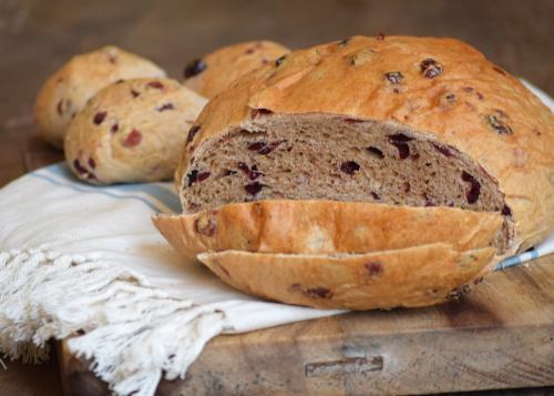 A loaf of bread with cranberries, sliced on a wooden board, with a few round rolls in the background.