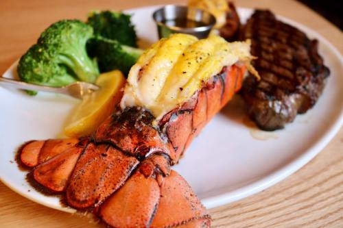 A plate featuring grilled lobster tail, steak, broccoli, and lemon, served with a small cup of sauce.