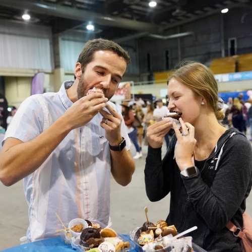 A man and woman enjoy ice cream treats at a crowded event, smiling as they take big bites.