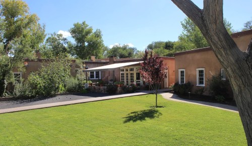 A charming adobe-style house surrounded by greenery and a well-maintained lawn under a clear blue sky.