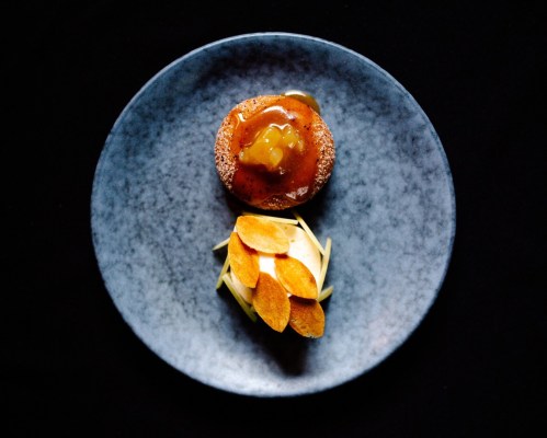 A dessert on a dark plate featuring a round pastry topped with caramel and orange slices beside a creamy base.