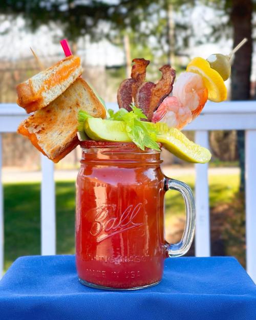 A mason jar filled with a Bloody Mary, garnished with shrimp, bacon, pickles, celery, and a grilled cheese sandwich.