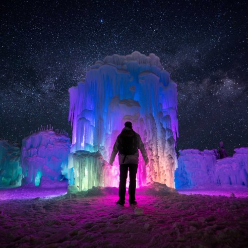 A person stands in an ice formation illuminated with colorful lights under a starry night sky.